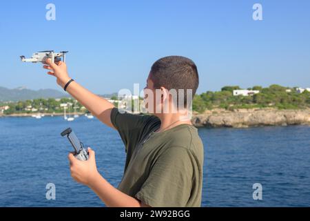 Heureux adolescent garçon, préparant et finalisant les détails du vol de drone pendant la journée ensoleillée avec la mer méditerranée en arrière-plan Espagne Banque D'Images