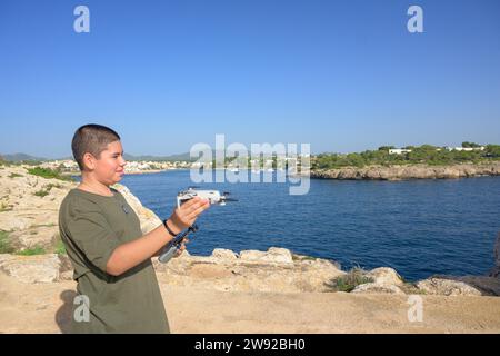 Heureux adolescent garçon, préparant et finalisant les détails du vol de drone pendant la journée ensoleillée avec la mer méditerranée en arrière-plan Espagne Banque D'Images