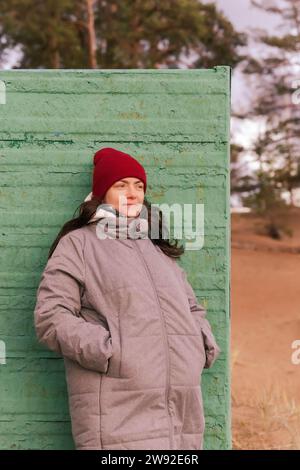 une femme portant une veste d'automne et un chapeau rouge marche le long d'une plage d'automne et pose à côté d'un vieux vestiaire Banque D'Images