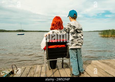 Une jeune fille aux cheveux roux, représentante de la génération Z, et son frère cadet sont assis sur une chaise sur la rive d'un lac tranquille, appréciant TH Banque D'Images