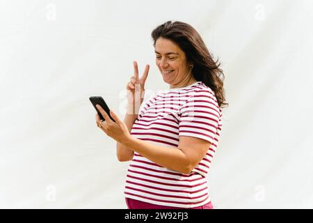 Une femme caucasienne aux cheveux longs, positive, d'âge moyen dans un chemisier rayé montre un signe de victoire tout en parlant sur un téléphone portable sur un fond clair Banque D'Images