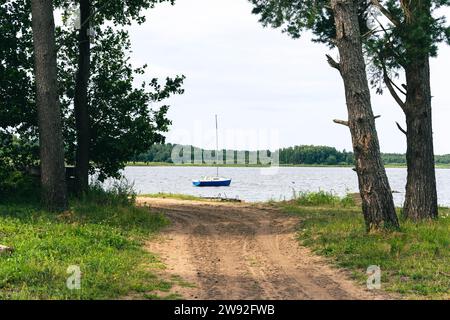 Une vue panoramique imprenable sur un petit voilier reposant paisiblement sur un lac serein, avec un charmant chemin de terre menant à l'eau, entouré par natur Banque D'Images