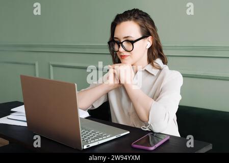 Connecté et souriant. Femme avec casque sans fil s'engage dans l'appel vidéo d'ordinateur portable avec joie. Conversation numérique. Femme en lunettes Smiles pendant Lapt Banque D'Images