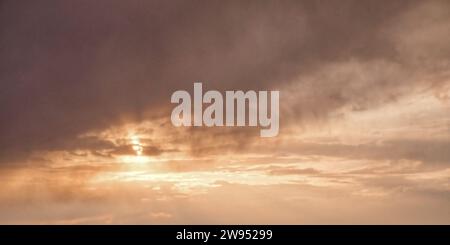 L'image montre un ciel nuageux avec le soleil jetant un regard à travers, projetant une lueur chaude au milieu des nuages sombres. Banque D'Images