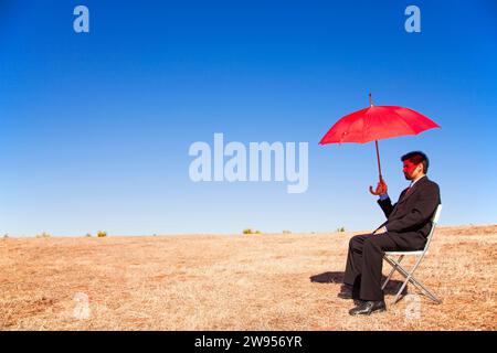 Businessman situés sur une chaise dans un paysage tenant un parapluie rouge Banque D'Images
