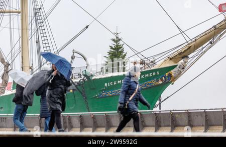 Hambourg, Allemagne. 24 décembre 2023. Les marcheurs passent devant le navire du musée Rickmer Rickmers sur l'Elbe dans la bruine. La tempête s'est affaiblie, mais beaucoup de pluie et de neige sont attendues dans certaines régions d'Allemagne ce week-end. Les niveaux d'eau de nombreuses rivières augmenteront également. Les voyageurs ferroviaires doivent être prêts pour les trains complets. Crédit : Markus Scholz/dpa/Alamy Live News Banque D'Images