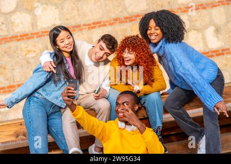 Cool groupe diversifié d'amis adolescents prenant un selfie ensemble assis sur un banc en bois à l'extérieur Banque D'Images