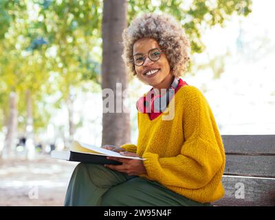 Portrait d'une femme afro-américaine souriante avec des lunettes et des cheveux afro regardant la caméra assis dans un parc tout en lisant un livre Banque D'Images