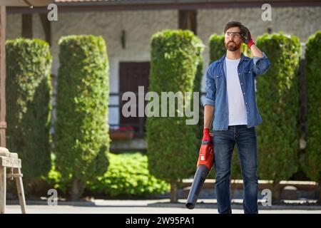 Bel homme à main caucasien debout avec souffleur à main, tout en ayant une pause après avoir travaillé à l'extérieur. Vue de face du jardinier barbu portant des lunettes de sécurité avant de nettoyer la cour arrière. Concept de lieu de travail. Banque D'Images