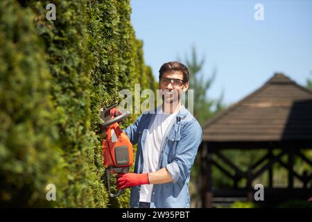 Jardinier professionnel taillant, façonnant la haie de thuja à feuilles persistantes avec un coupe-herbe électrique moderne à l'heure d'été. Portrait de travailleur souriant regardant la caméra, tout en élaguant. Concept de services de jardinage. Banque D'Images