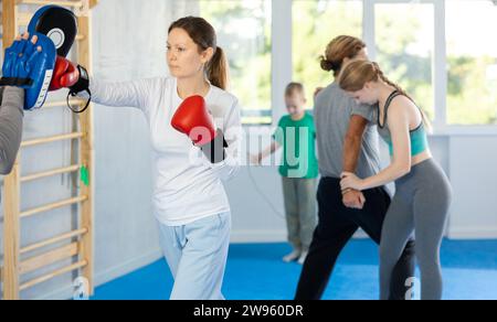 Femme dans les gants de boxe pratique des coups de poing avec entraîneur dans les mitaines dans le club de sport pendant l'entraînement familial Banque D'Images