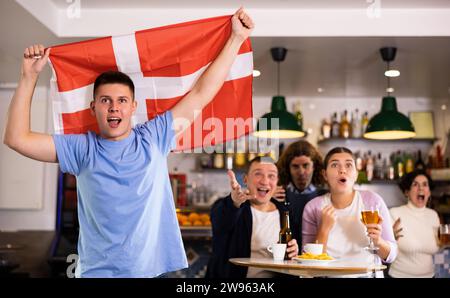 Crier les jeunes fans de sport pour adultes qui se sont pris pour l'équipe préférée et qui ont le drapeau du Danemark tout en regardant le match ensemble au pub Banque D'Images