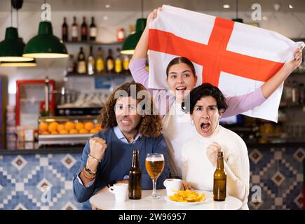 Groupe d'amis, supporters sportifs acclamant l'équipe favorite avec drapeau de l'Angleterre tout en regardant le match à la télévision dans le bar Banque D'Images