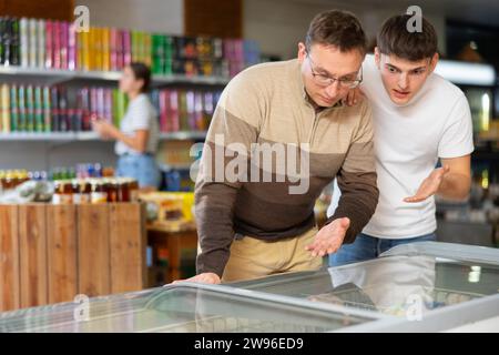 L'homme avec des lunettes et le jeune gars choisissent ensemble des aliments congelés dans le réfrigérateur dans le supermarché. Photo de haute qualité Banque D'Images