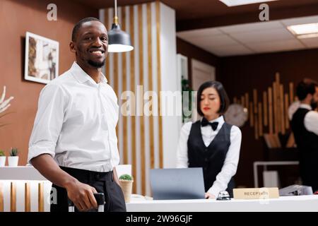 Touriste masculin assis à la réception pour s'inscrire et remplir les formulaires d'enregistrement, client souriant et attendant le service de l'hôtel. Jeune homme debout près du comptoir de réception dans le hall, complexe de luxe. Banque D'Images