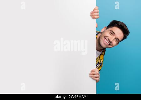 Photo de jeune drôle gars heureux cachant la bannière blanche d'information de panneau d'affichage de mur avec des détails promo isolé sur fond de couleur bleue Banque D'Images