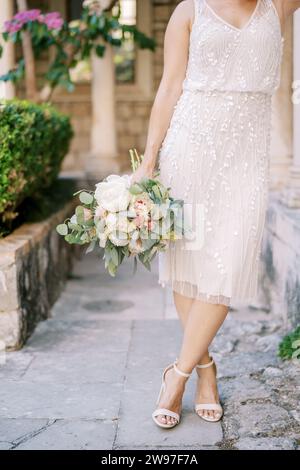 Mariée avec un bouquet de fleurs se tient les pattes croisées sur les pavés dans le jardin. Rogné. Sans visage Banque D'Images