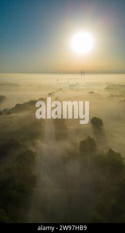 Cette image atmosphérique capture une vue aérienne à couper le souffle d'une forêt dense recouverte de brume lors d'un lever de soleil matinal. Le soleil, positionné au centre du cadre, projette une lueur chaude sur le paysage brumeux, créant une scène sereine et presque éthérée. La cime des arbres émerge du brouillard, suggérant l'immensité de la forêt et la beauté des moments de la nature qui sont souvent invisibles depuis le sol. Misty Sunrise, vue aérienne sur la forêt. Photo de haute qualité Banque D'Images