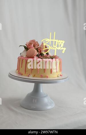 glaçage givré frais égouttant gâteau d'anniversaire rose vanille vraies roses et décoration macaron sur le dessus avec signe doré Banque D'Images