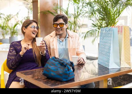 Heureux couple indien assis dans la cafétéria du centre commercial profitant avec tasse de café à la main et sac en papier, sac à main sur la table. Banque D'Images