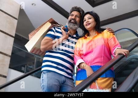 Heureux couple indien tenant sur la main courante de l'escalator et l'escalier roulant dans le centre commercial avec des sacs en papier, achats dans les mains. Concept shopaholic. Banque D'Images