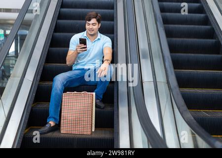 Heureux jeune homme indien assis, monter escalator et en utilisant un téléphone intelligent dans un centre commercial avec des sacs en papier, des achats. Concept shopaholic. Banque D'Images