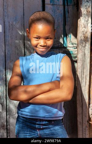 enfant africain debout avec les bras croisés dans la porte de sa cabane, maison de boue, dans un village informel, décontracté habillé de denim et t-shirt bleu Banque D'Images