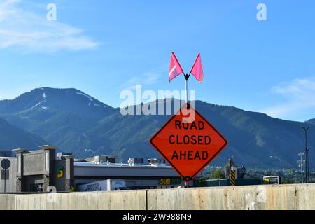 Panneau d'avertissement orange « ROUTE FERMÉE DEVANT » avec deux petits drapeaux sur le dessus avec des montagnes en arrière-plan Banque D'Images