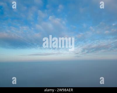 Cette photographie, prise avec un drone au-dessus de la brume, présente une vue à couper le souffle semblable à être au-dessus du niveau des nuages. La brume couvre le paysage, créant un horizon sans couture où le ciel se confond avec la terre dans une étendue bleue tranquille. Le ciel est orné de délicats nuages d'altocumulus, teintés des doux pastels de l'aube, suggérant le début en douceur d'une nouvelle journée. L'image donne un sentiment de paix et d'isolement, haut au-dessus du monde obscurci et éveillé ci-dessous. Au-dessus des nuages : vue aérienne d'un paysage recouvert de brume à l'aube. Photo de haute qualité Banque D'Images