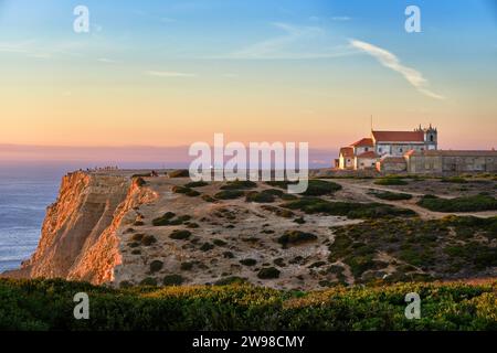Belle vue sur le vieux monastère historique sur le bord de la falaise pendant l'heure dorée du coucher du soleil. Océan calme, ciel coloré, certaines personnes apprécient la vue tranquille de l'été, coucher du soleil, ciel clair, quelques nuages, terrain accidenté de paysage rocheux. Portugal. Banque D'Images