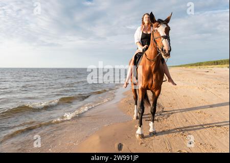 Une jeune femme chevauchant un cheval brun le long du bord de mer par une journée ensoleillée Banque D'Images