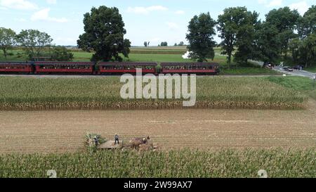 Une vue aérienne de six chevaux tirant une machine de récolte de maïs Amish, alors que le train de passagers à vapeur traverse la campagne Amish en automne Banque D'Images