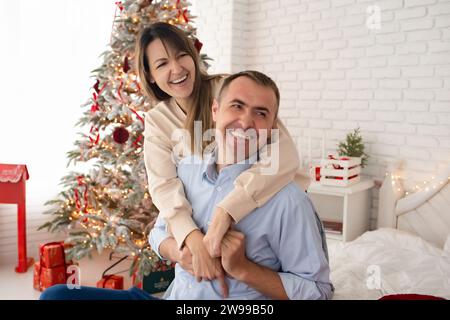 Un homme et une femme se sont embrassés et sont assis sur un lit dans une grande et belle pièce décorée pour Noël. Famille près de l'arbre du nouvel an dans une pièce lumineuse Banque D'Images