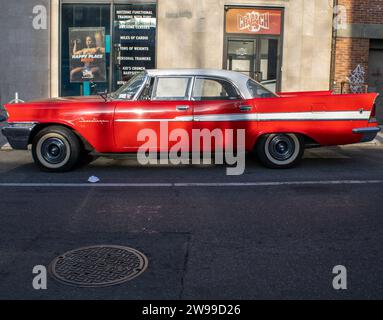 Une voiture rouge vintage est garée devant un bâtiment en briques à plusieurs étages, avec un homme mature silhouetté sur fond d'architecture Banque D'Images