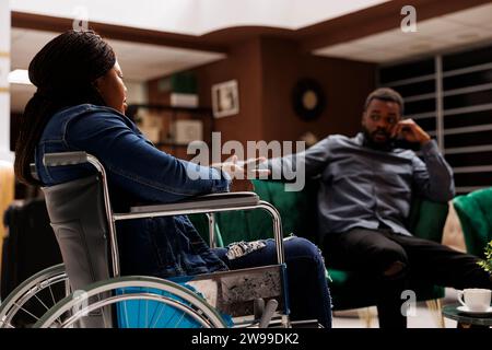 Jeune femme afro-américaine touriste avec handicap assis dans le hall de l'hôtel avec mari, arrivant à l'hôtel accessible en fauteuil roulant. Homme valide voyageant avec une petite amie handicapée Banque D'Images