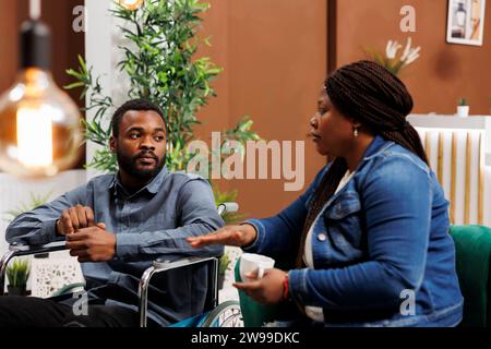 Couple afro-américain arrivant au centre de réadaptation pour handicapés, jeune homme utilisateur de fauteuil roulant voyageant avec sa femme, assis ensemble dans le hall de l'hôtel attendant une chambre avec accès handicapés Banque D'Images