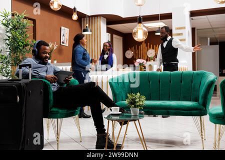 Souriant homme afro-américain voyageur utilisant une tablette dans le salon de l'hôtel, touriste noir portant des écouteurs assis avec valise dans le hall écoutant de la musique en attendant le processus d'enregistrement Banque D'Images