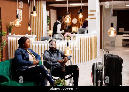 Jeune Afro-américain handicapé utilisateur de fauteuil roulant voyageant avec un assistant personnel, un handicapé et sa femme assis dans le hall de l'hôtel avec des bagages. Expérience client pour les personnes handicapées Banque D'Images