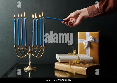 Main de jeune femme méconnaissable allumant des bougies sur Hanukkah menorah debout sur la table à côté de la pile de boîtes-cadeaux avec des cadeaux Banque D'Images