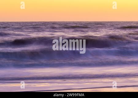 Deux personnes portant des combinaisons et portant des planches marchent le long d'une plage de sable au crépuscule alors que le soleil se couche en arrière-plan Banque D'Images