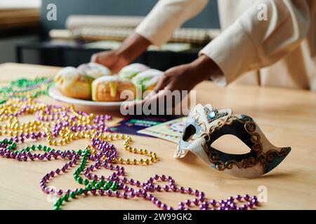 Masque de carnaval et perles colorées couchées sur la table en bois tandis que jeune femme mettant assiette avec des beignets traditionnels faits maison Banque D'Images