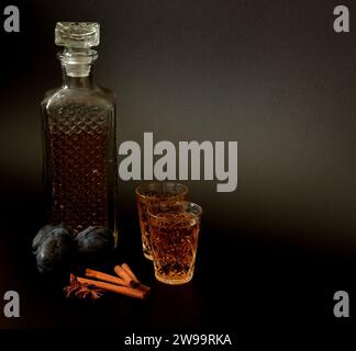 Liqueur de prune dans une carafe en cristal et deux verres sur un fond noir, à côté de fruits mûrs et cannelle à l'anis. Gros plan. Banque D'Images