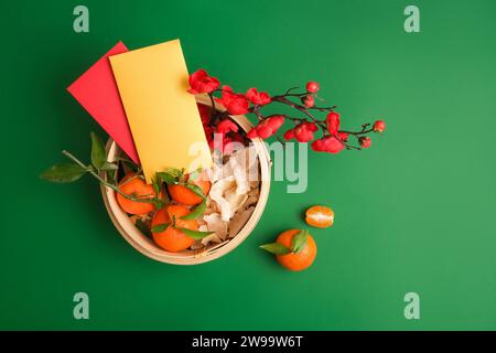 Mandarines, confiture de gingembre, branches de fleurs de pêche et enveloppes porte-bonheur à l'intérieur d'un cuiseur vapeur en bambou sur fond vert. Espace libre pour la conception. Banque D'Images