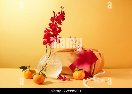 Un vase d'orchidées rouges, une boîte cadeau enveloppée de soie beige, des mandarines et une ficelle de perles se détachent sur le fond jaune. Espace publicitaire. Banque D'Images