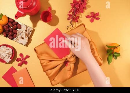 Vue de dessus d'une main remplissant une enveloppe rouge dans un sac cadeau. Un service à thé, des orchidées rouges, des mandarines, des jujubes, de la confiture et des enveloppes porte-bonheur sont arra Banque D'Images
