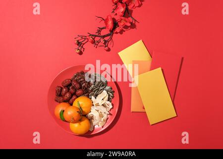 Mandarines, confiture de gingembre, jujube, graines de melon et graines de tournesol sont servis sur une assiette, enveloppes porte-bonheur et branche de fleur de pêche sur un dos rouge Banque D'Images