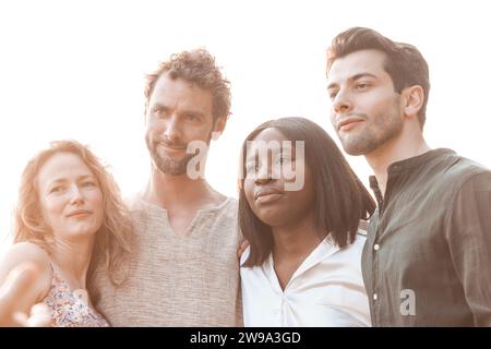dans la lueur tendre du soleil de l'après-midi, un groupe diversifié d'amis se tient près les uns des autres, incarnant l'esprit d'unité. La photographie capture un homme et une femme caucasiens, une femme noire et un homme du Moyen-Orient, partageant tous un moment de connexion et de joie tranquille, leurs visages se dormant dans la douce lumière du soleil. Sunlit Bonding : divers amis en harmonie. Photo de haute qualité Banque D'Images