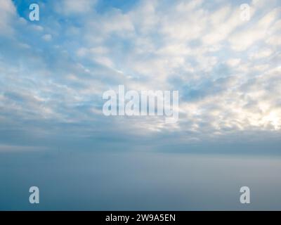 Cette image, capturée par un drone au-dessus de la brume, représente un paysage de ciel serein où le blanc doux des nuages d'altocumulus rencontre le doux gradient de la lumière de l'aube. La couverture de brume crée un horizon minimaliste, mêlant ciel et terre en une étendue harmonieuse de bleus et de blancs sourds. Le soleil, partiellement caché, apporte un léger contre-jour aux nuages, suggérant le réveil tranquille de la journée. Cette scène résume la tranquillité et l'immensité du monde naturel, intact et expansif. Skyscape serein au-dessus de la brume enveloppait Horizon. Photo de haute qualité Banque D'Images