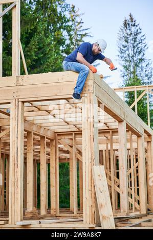 Menuisier construisant en bois, maison encadrée de deux étages près de la forêt. Homme barbu portant des lunettes clouant l'ongle avec casque de protection. Le concept de construction écologique moderne. Banque D'Images