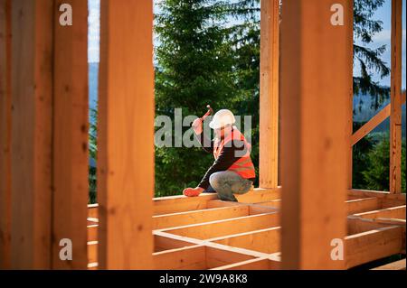 Carpenter construisant une maison en bois de deux étages près de la forêt. Homme martelant des clous dans la structure, portant un casque de protection et un gilet de construction. Concept de construction écologique moderne. Banque D'Images